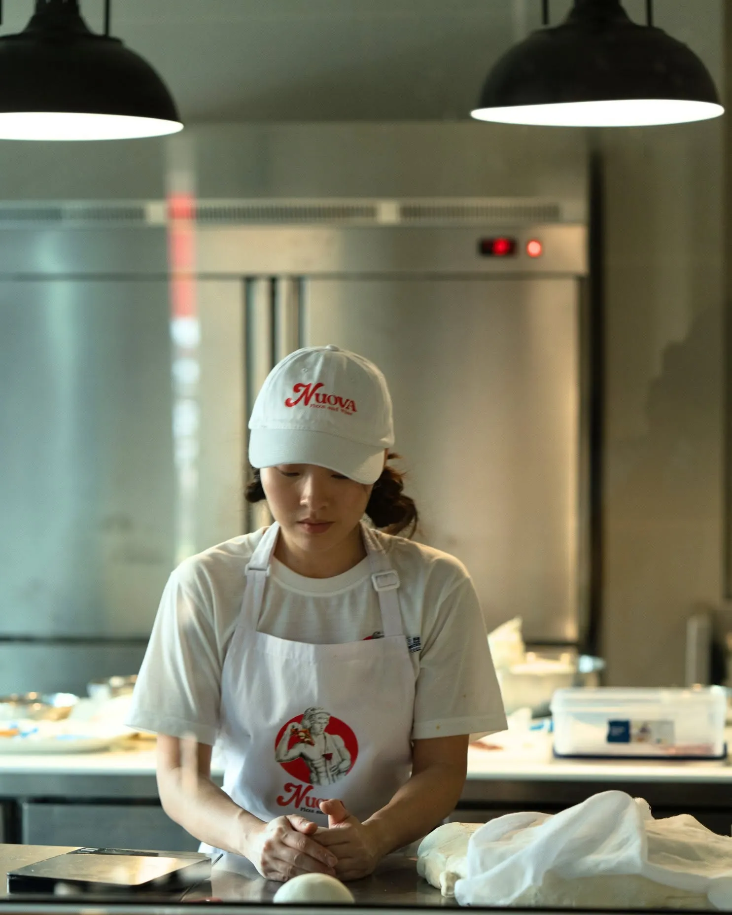 The chef in a Nuova cap and logo apron at the steel bench, a dough ball ready in front of him